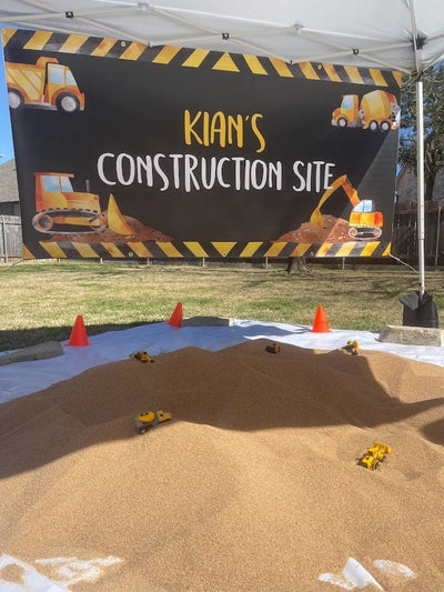 A construction themed birthday party with a black banner that says &#39;Kian&#39;s Construction Site&#39; with yellow construction vehicles. There is a sandbox with yellow toy construction vehicles in it.