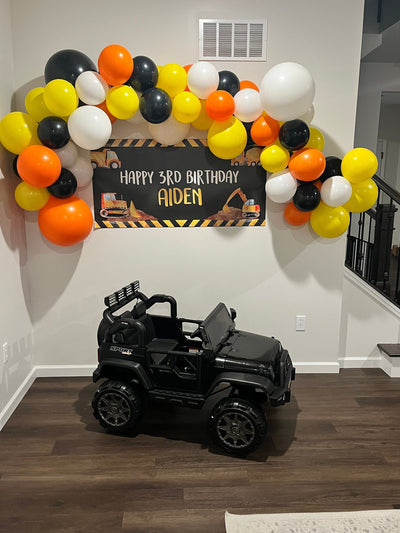 a construction birthday banner with happy birthday writing and construction vehicles featuring a balloon garland surrounding the banner. A kids ride on jeep below