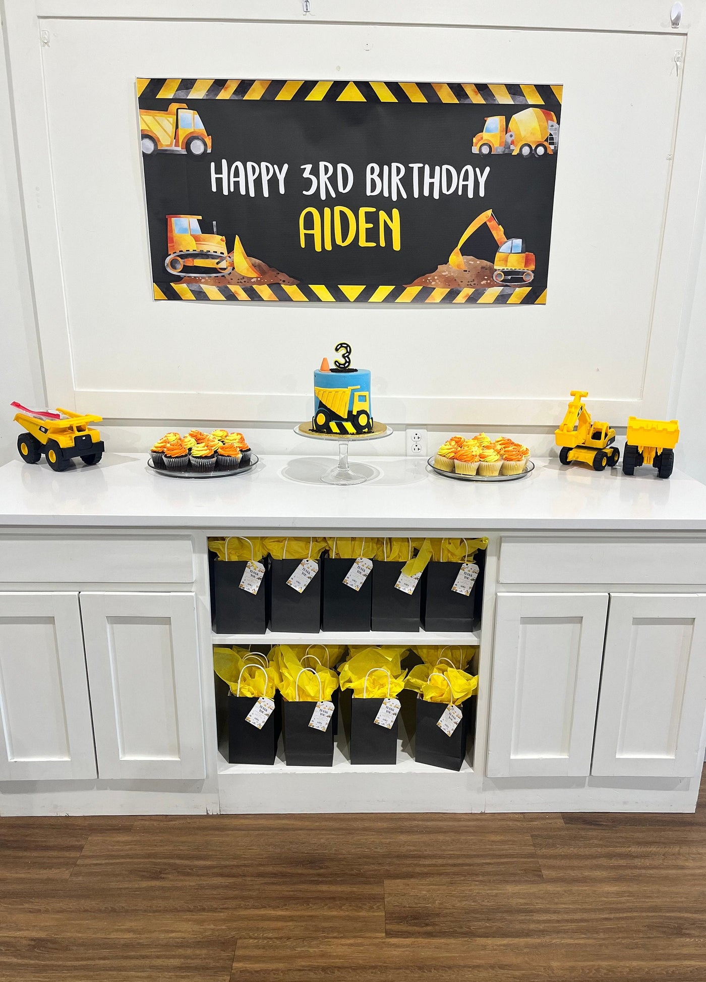 A white table with a black and yellow construction themed personalized banner that reads &#39;Happy Birthday&#39;. There are cupcakes with yellow and orange frosting, a yellow and blue construction themed cake, and yellow construction toys.