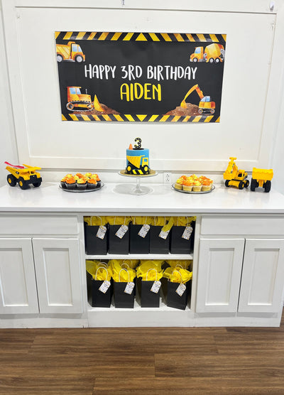 A white table with a black and yellow construction themed personalized banner that reads &#39;Happy Birthday&#39;. There are cupcakes with yellow and orange frosting, a yellow and blue construction themed cake, and yellow construction toys.