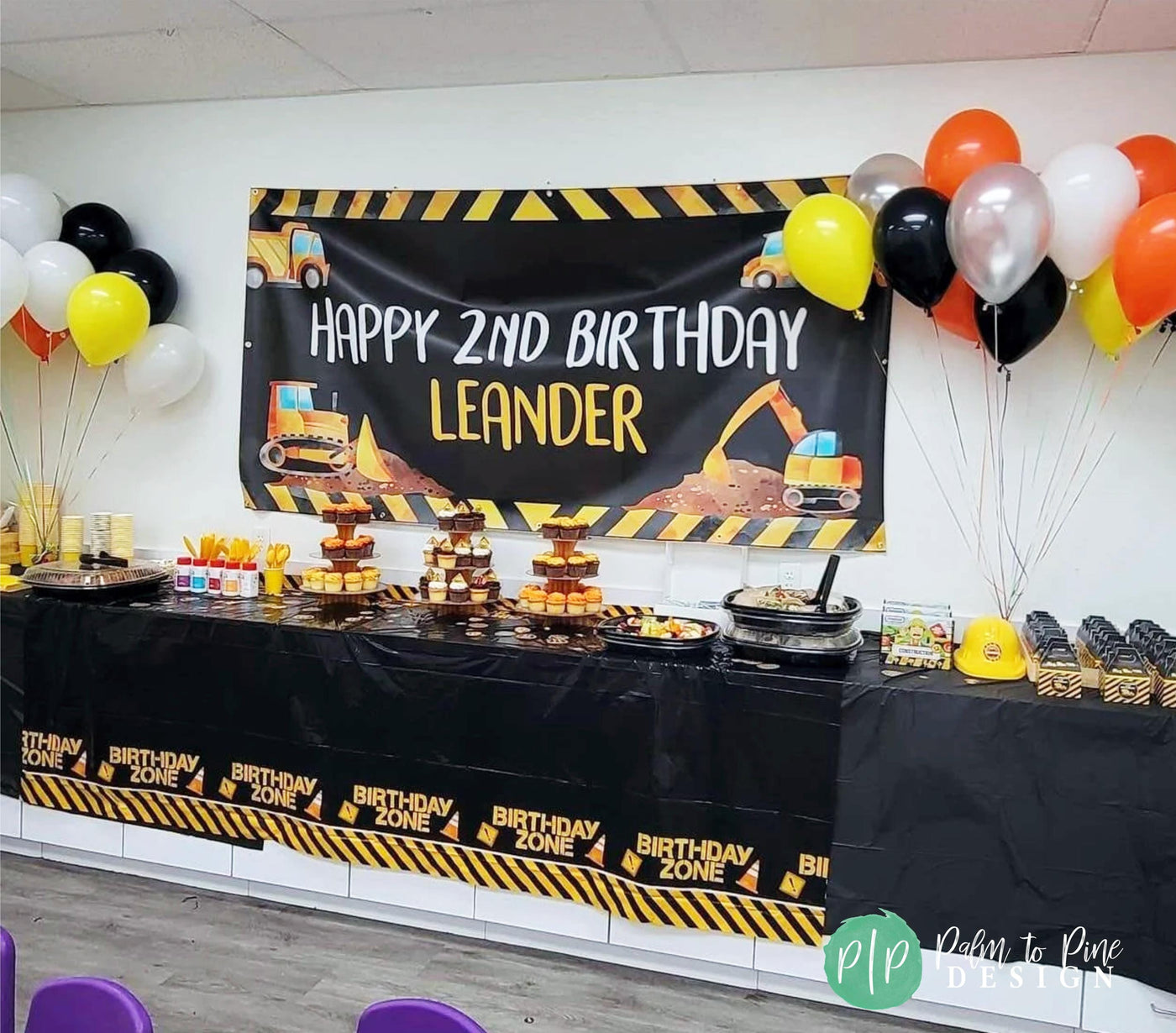 A black table with a yellow and black construction themed tablecloth. The table is decorated with cupcakes, snacks, and a banner that reads &#39;Happy Birthday Name.&#39; There are yellow, orange, and silver balloons in the background.